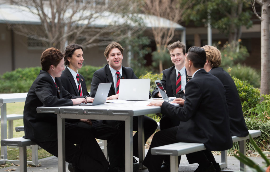 A group of students laugh together as they work on their laptops at an outdoor table